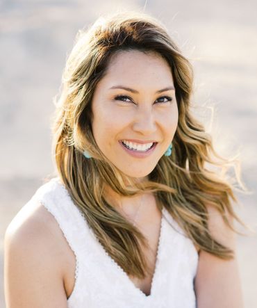 Woman smiling, wearing a white dress, turquoise earrings, and wavy blonde hair against a blurred light background.