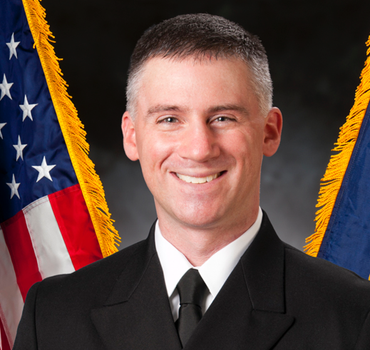 Man in navy uniform smiles in front of flags.