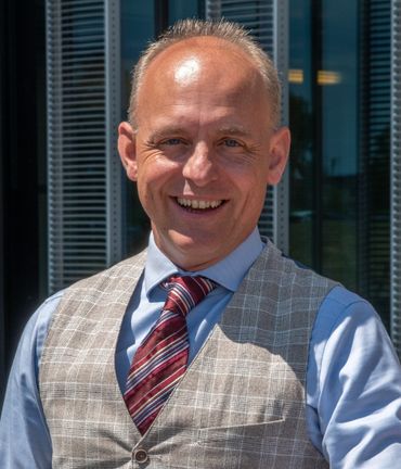 Man smiling, wearing a vest and tie, standing outside a modern building.