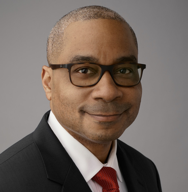Man in a suit, wearing glasses, smiling slightly, against a gray background.