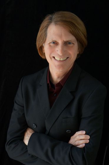 Man with brown hair, crossed arms, wearing a black blazer and burgundy shirt, smiling against a black background.