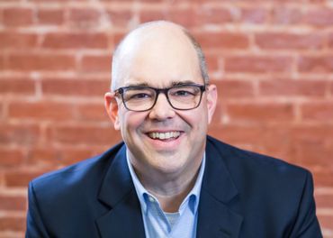 Man with glasses smiles, wearing a blue shirt and blazer against a red brick wall.