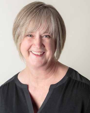 Woman in a dark suit, curly brown hair with blonde highlights, hand to chin, smiling.