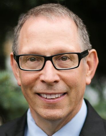 Man in black glasses smiles, wearing a suit jacket and light blue shirt.