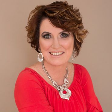 Woman with short, curly auburn hair, wearing a red top and silver jewelry, smiles at the camera.