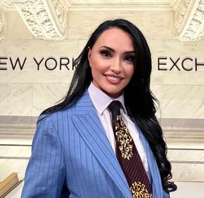 Woman in blue pinstripe suit, standing in front of the New York Stock Exchange sign, smiling.
