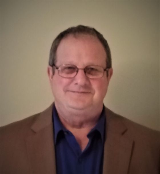 Man wearing glasses, brown blazer, and blue shirt, smiling.