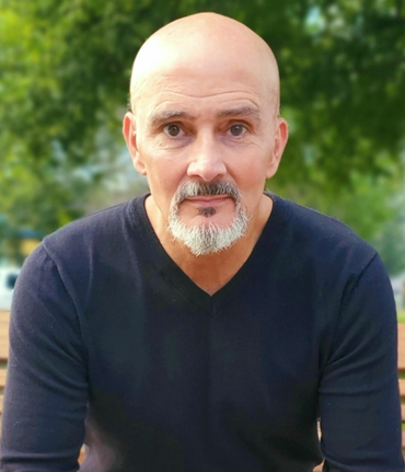 Bald man with a grey goatee and a serious expression, wearing a black v-neck shirt, seated outside.