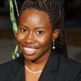 Woman with braided hair, smiling, wearing a black blazer and pearl necklace.