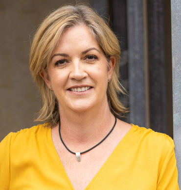 Woman with blonde hair wearing a yellow top and black necklace smiles in an outdoor setting.