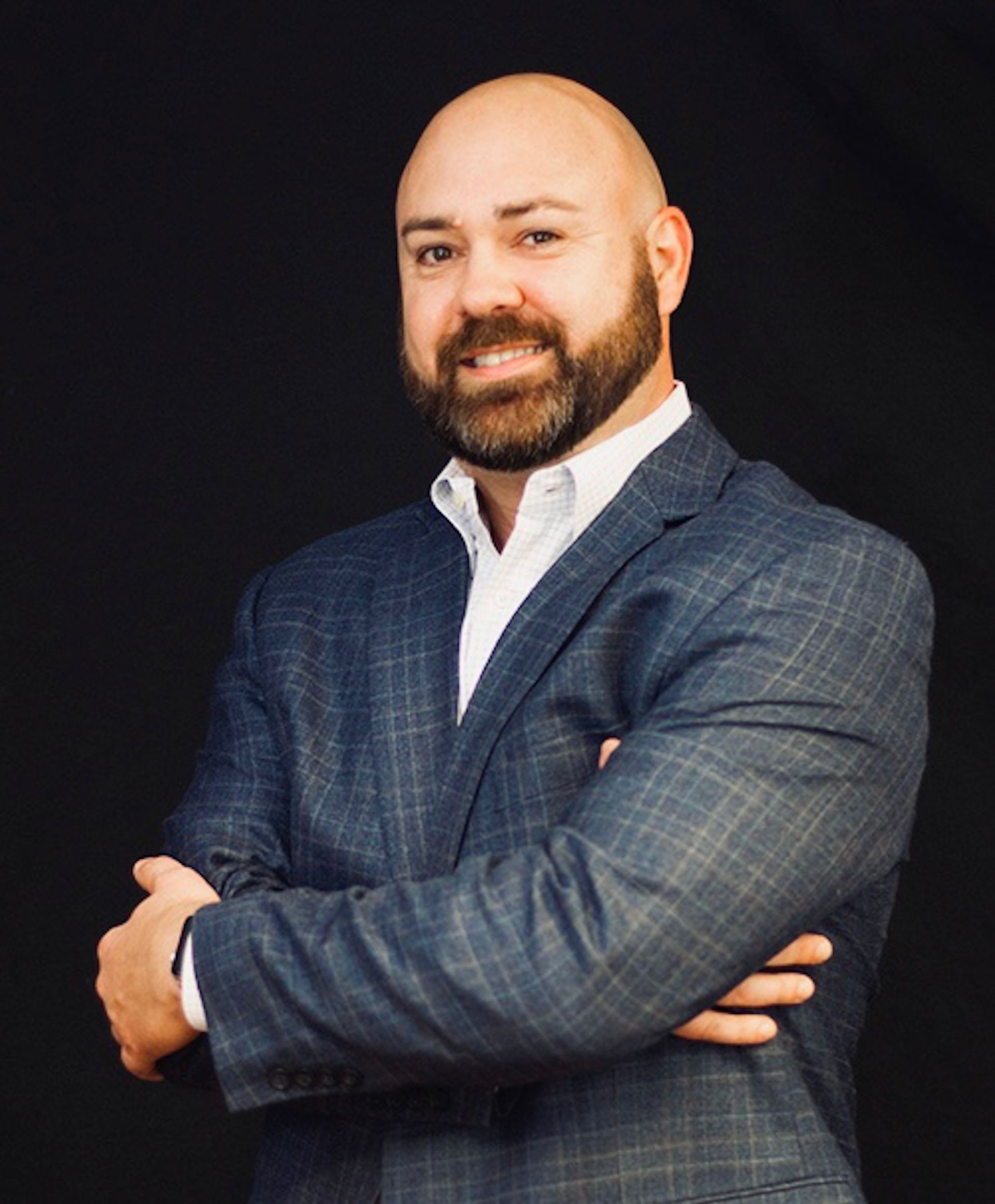 Bald man in a blue plaid suit and open white shirt, arms crossed, smiling, black background.