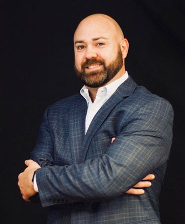 Bald man in a blue plaid suit and open white shirt, arms crossed, smiling, black background.