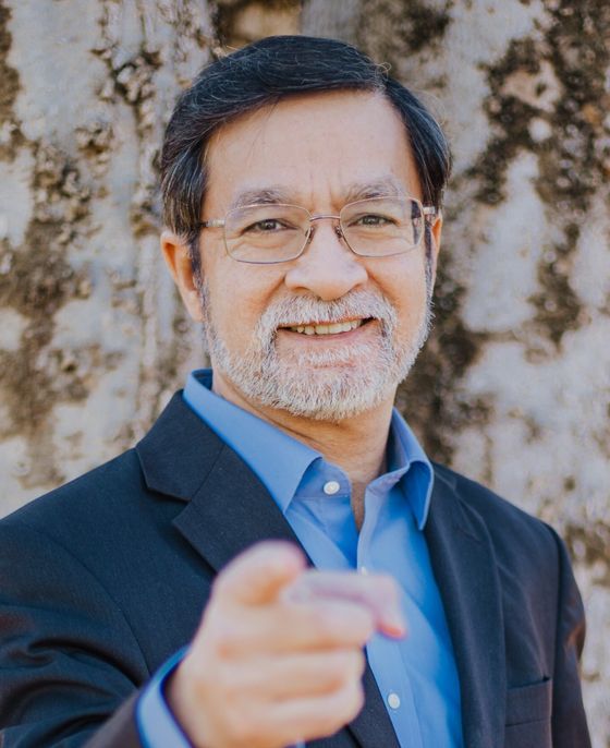 Man with glasses points, smiling, wearing blue shirt and suit jacket, against a stone wall.