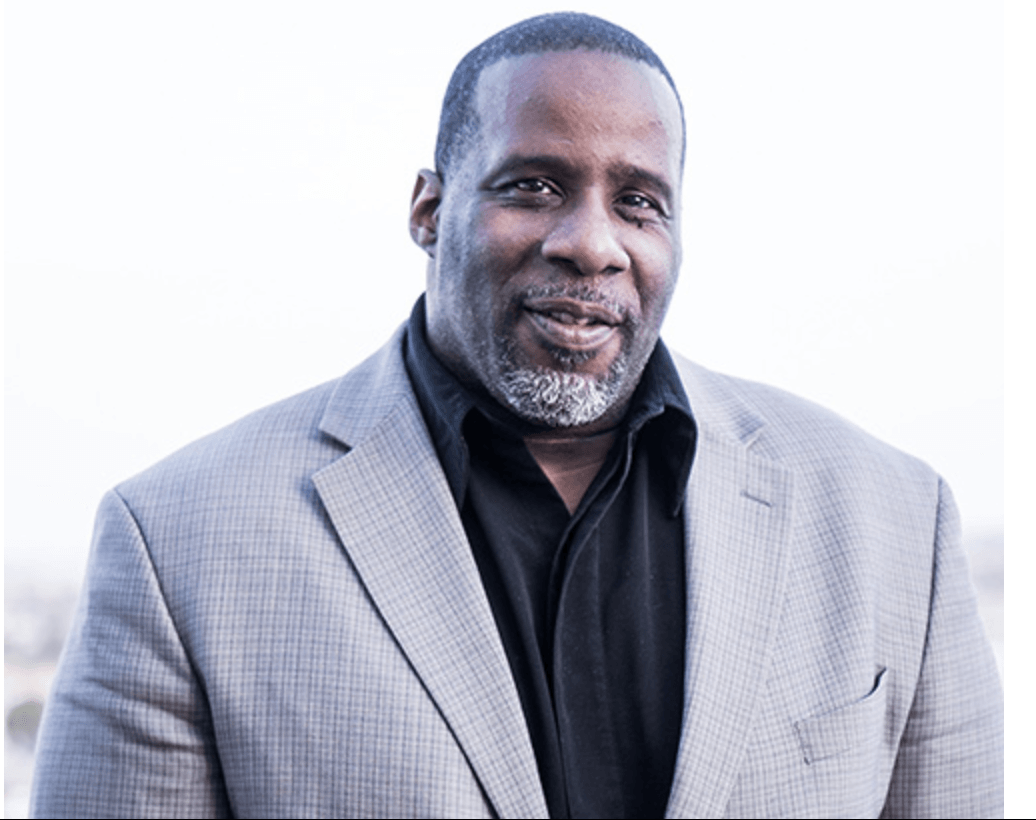 Man in gray blazer and black shirt smiles outdoors.