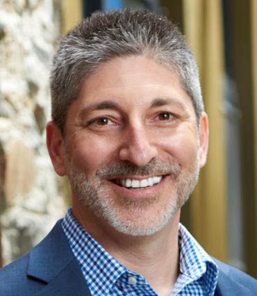 Man wearing glasses and a tweed cap, smiling in a hallway.