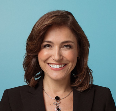 Woman with brown hair smiling, wearing a black blazer and necklace, against a blue background.