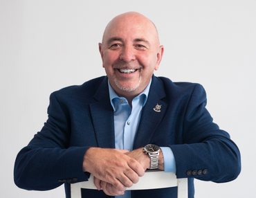 Bald man in blue suit smiles, leans on white chair, silver watch.