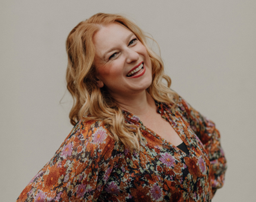 Woman with wavy auburn hair smiles widely, leaning to her left. She wears a floral print top, standing in front of a neutral backdrop.