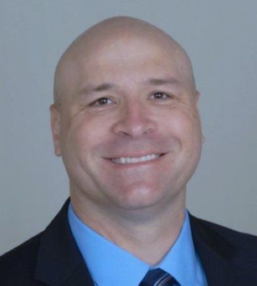 Bald man smiling in a suit and light blue shirt, against a neutral background.