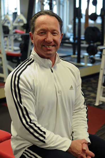 Man in a white Adidas zip-up jacket smiles in a gym setting, arms crossed, with workout equipment visible.