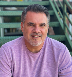 Man with graying hair smiles, wearing a purple shirt, posing outdoors near a metal staircase.