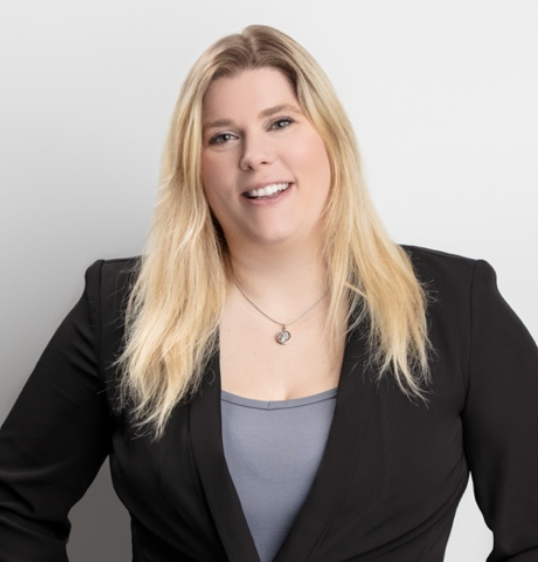 Woman with blonde hair, smiling, wearing a black blazer and gray top, posing in front of a white background.