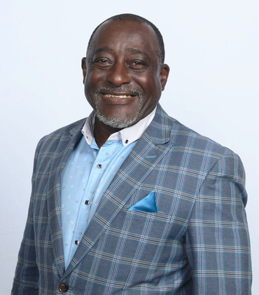 Smiling man in a checkered blue blazer, light blue shirt, and a blue pocket square; white backdrop.
