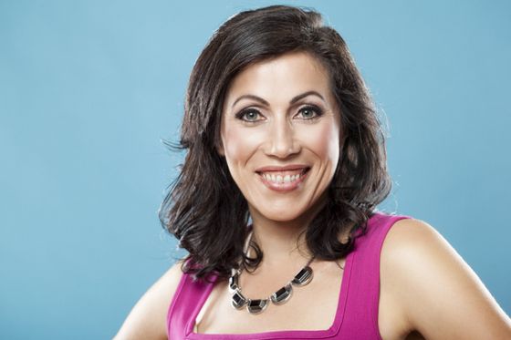 Woman with dark hair smiles, wearing pink dress and necklace against a blue backdrop.