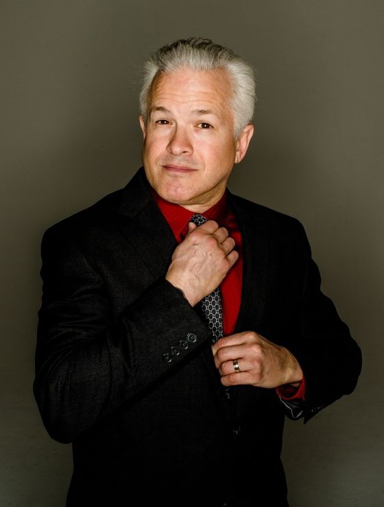 Man in suit, adjusting tie, looking at the camera against a gray background.