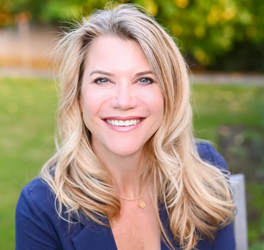 Blond woman smiles at camera, wearing a blue blazer and gold necklace, seated outdoors with a green background.