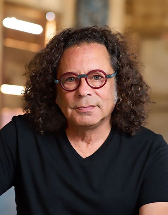 Man with curly hair and round glasses, wearing a black shirt, indoors with a neutral expression.