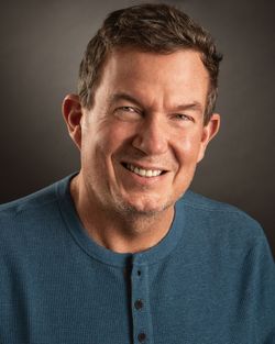 Man with a warm smile, wearing a blue Henley sweater, against a dark background.