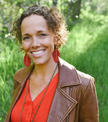 Woman smiling outdoors, wearing red shirt, brown leather jacket, and red feather earrings.