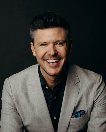 Man in a light gray blazer, smiling with a dark patterned shirt, dark background.