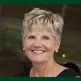 Blonde woman smiling, wearing pearl earrings, black top, outdoors with blurred background.