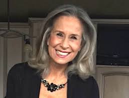 Woman with gray hair smiles, wearing a black top and a jeweled necklace. Indoors, neutral background.