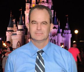 Man in blue shirt and tie, smiling, in front of Cinderella Castle at night with people in the background.