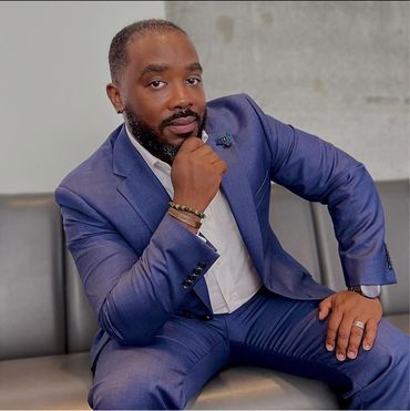 Man in blue suit, resting chin on hand, seated on a gray sofa in front of a concrete wall.