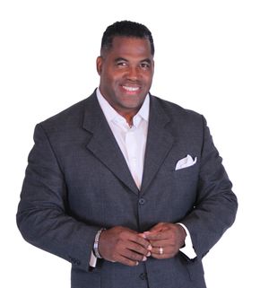 Man in gray suit and white shirt, smiling, hands clasped. Pocket square visible.