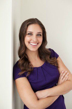 Woman with wavy brown hair smiles, arms crossed, wearing a purple dress, leaning against a white wall.