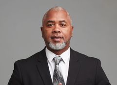 Man in a black suit with a patterned tie; he has gray hair and beard, and is looking at the camera.