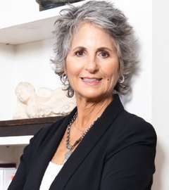 Woman with gray curly hair, wearing a black blazer and necklace, smiles at the camera, standing in front of a shelf.