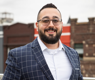 Man in a blue plaid blazer smiles outdoors, with glasses and a beard. Buildings in the background.