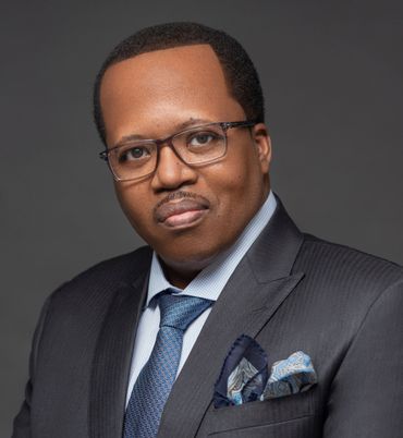 Man in suit and glasses looking at the camera, wearing a patterned tie and pocket square, with a gray backdrop.