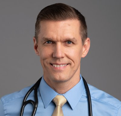 Doctor with stethoscope smiling, wearing a light blue shirt and gold tie.