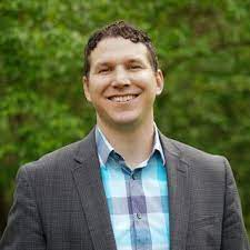 Man in a plaid shirt and gray blazer smiles in front of green foliage.