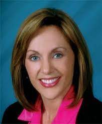 Woman with blonde hair, smiling, wearing a pink shirt and black jacket.