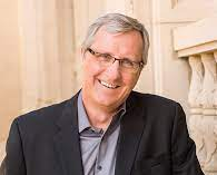 Man in glasses, smiling, wearing a dark suit jacket and gray shirt, in a light-colored building.