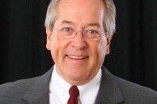 Man wearing glasses and a suit, smiling. Red tie, white shirt, dark background.