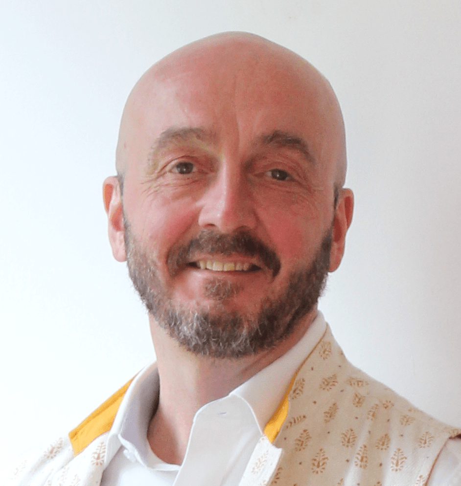 Smiling bald man with a beard, wearing a white shirt and patterned vest, indoors.